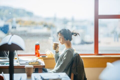 Student sitting at a table by a window, holding a hot beverage and wearing headphones.