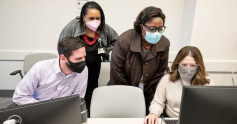 Four people wearing masks, two standing behind two who are seated on a computer.