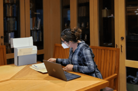 Archivist sitting at a table in the UCSF Archives Reading Room looking at documents with an open hollinger box to her right.