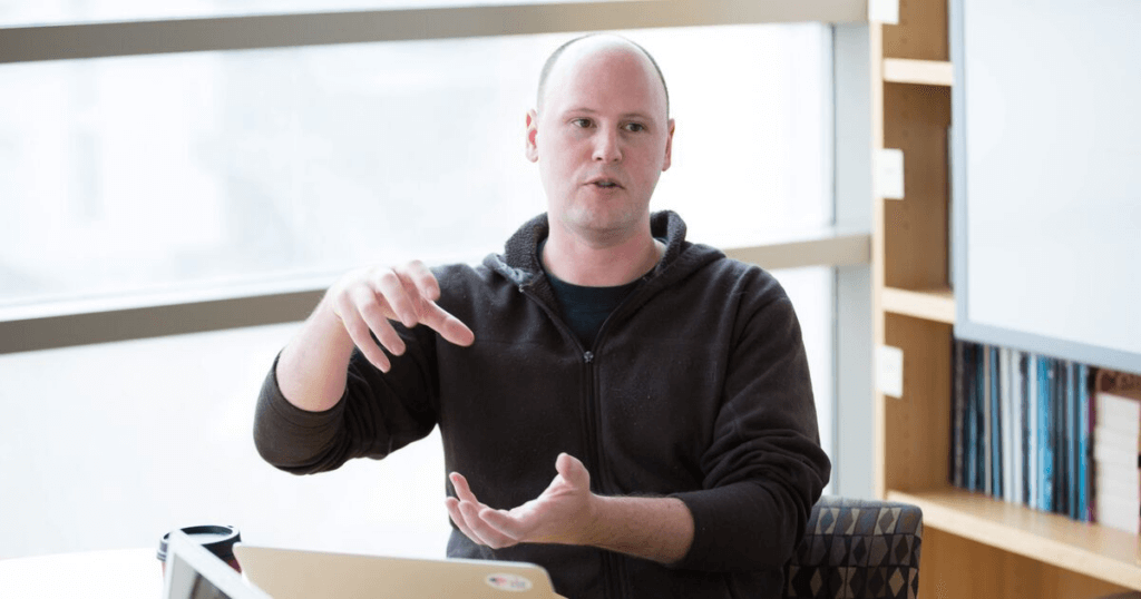 A man sitting at a table and gesturing with both hands while explaining information in a workshop.